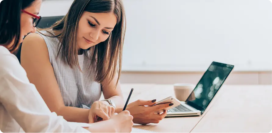 Two women collaborating at a table, one holding a smartphone and the other taking notes, with a laptop and coffee cup in the background, reflecting a professional setting for discussing marketing strategies.