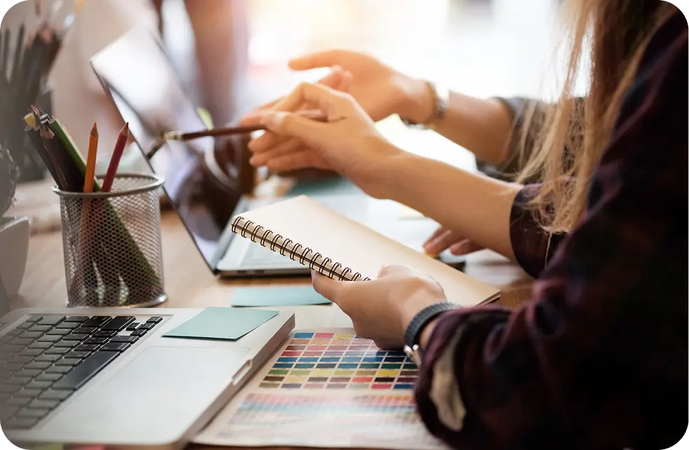 Hands collaborating over a laptop and notebook, discussing marketing strategies for recovery centers, with colorful design swatches and stationery in view.